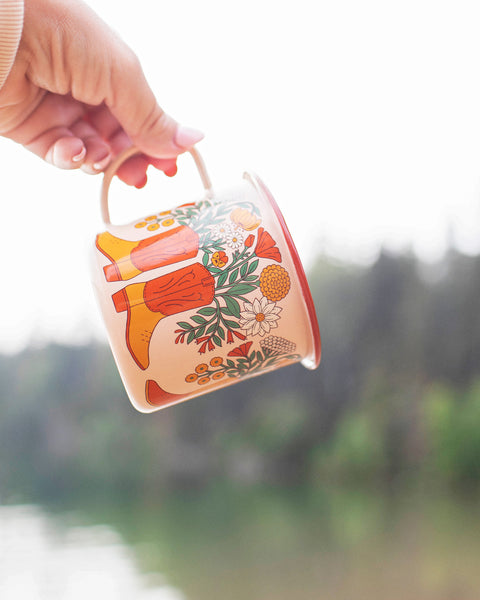Western Bloom Enamel Mug 16oz being held outdoors, showcasing vibrant floral and cowboy boot design.
