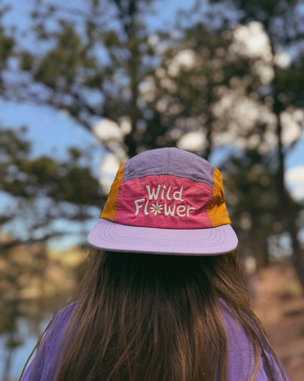 Child wearing a colorful hat with the text 'Wild Flower Kids Hat' against a natural backdrop.