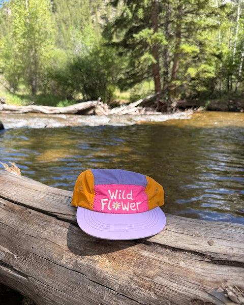 Wild Flower Kids Hat placed on a log by a peaceful stream surrounded by trees.