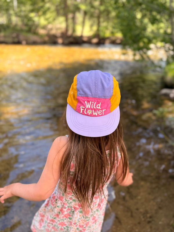 Child wearing a colorful Wild Flower Kids Hat by a river, highlighting style and fun for outdoor adventures.