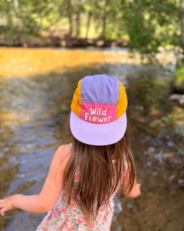 Child wearing a colorful Wild Flower Kids Hat by a river, highlighting style and fun for outdoor adventures.
