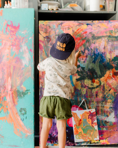 Child creating art while wearing a Wild Thing Kids Hat in a colorful studio background.