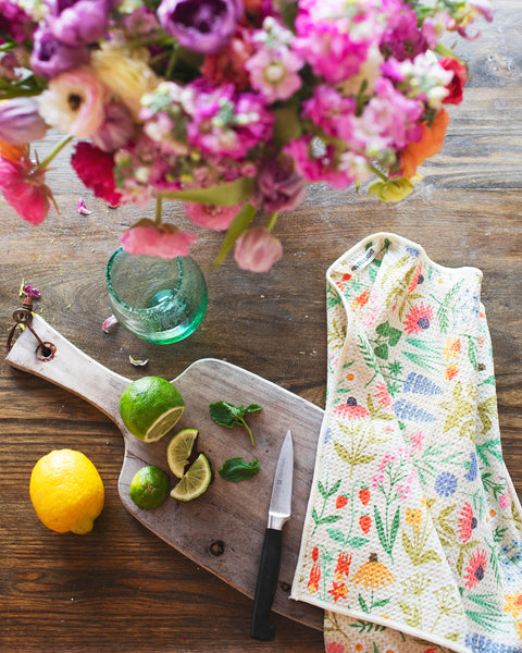 Wildflower Wonder Kitchen & Tea Towel next to sliced limes, lemons, and a bouquet of flowers on a wooden table.