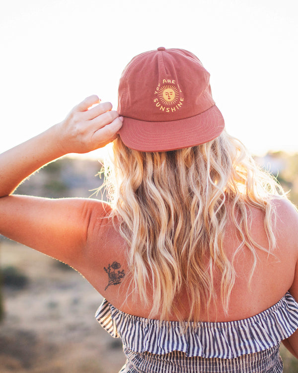 The You Are My Sunshine Hat in rust red with a yellow sun print, worn by a blonde woman seen from behind outdoors at sunset.