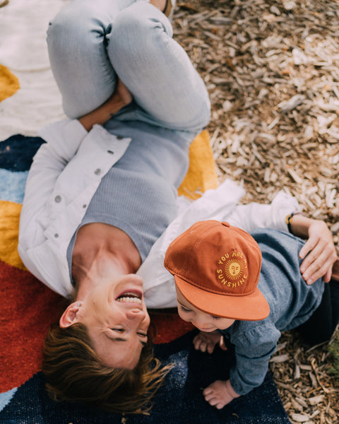 A happy child wearing a You Are My Sunshine Kids Hat, playing with a parent on a colorful blanket outdoors.