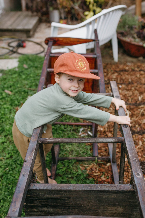 Child wearing Trek Light 'You Are My Sunshine' hat, playing outdoors with a rustic wooden frame, embodying adventure and joy.