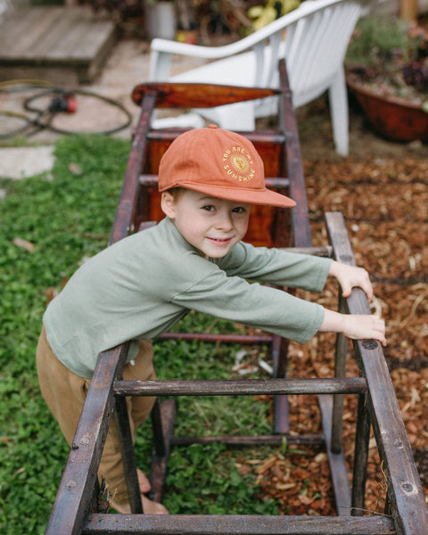 Child wearing Trek Light 'You Are My Sunshine' hat, playing outdoors with a rustic wooden frame, embodying adventure and joy.