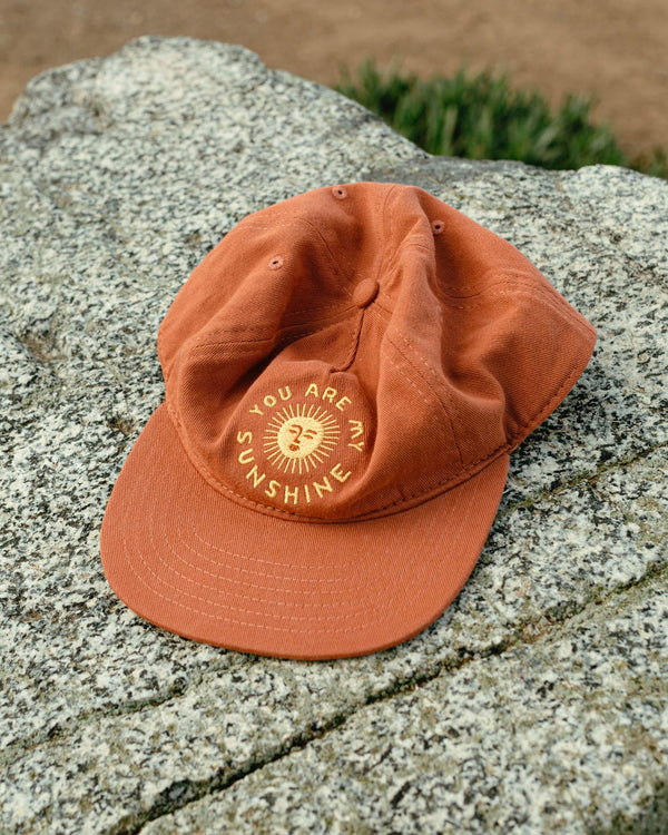 "You Are My Sunshine" hat in rust color on rock, part of matching hats set for kids and adults, perfect for Twinsie adventures. By Trek Light.