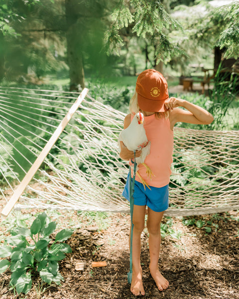 Child wearing a You Are My Sunshine Kids Hat, holding a chicken in a garden setting.
