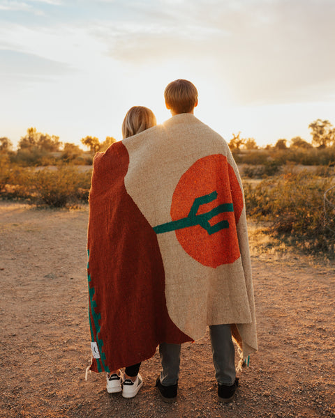 Couple wrapped in a cozy handwoven Arizona Blanket, made from recycled materials, enjoying a sunset in the desert. By Trek Light.