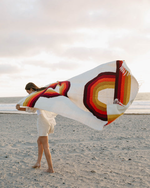 Person holding vintage rainbow Bowie Blanket on a beach, showcasing eco-friendly Trek Light design, handwoven with recycled materials.