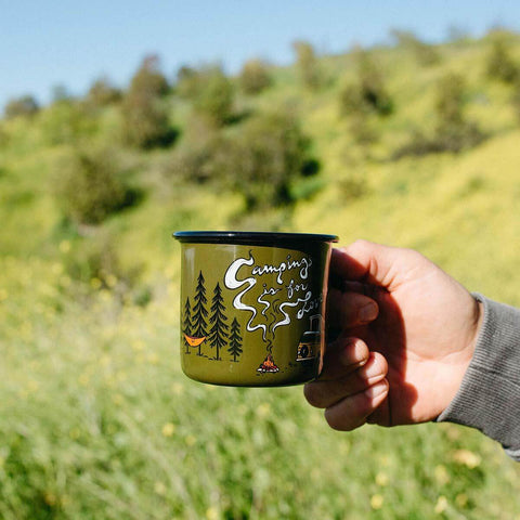 Trek Light 'Camping Is For Lovers' 12oz enamel mug in hand against a scenic outdoor backdrop, perfect for adventures and cozy sips.