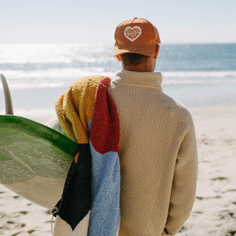 Person wearing 'Camping Is For Lovers' hat holds surfboard and colorful towel by the beach, representing Trek Light spirit.