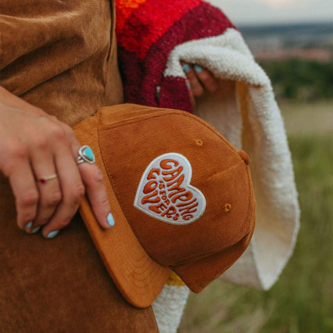 Person holding a 'Camping Is For Lovers' hat by Trek Light, perfect for outdoor adventures with a curved brim and snapback style.