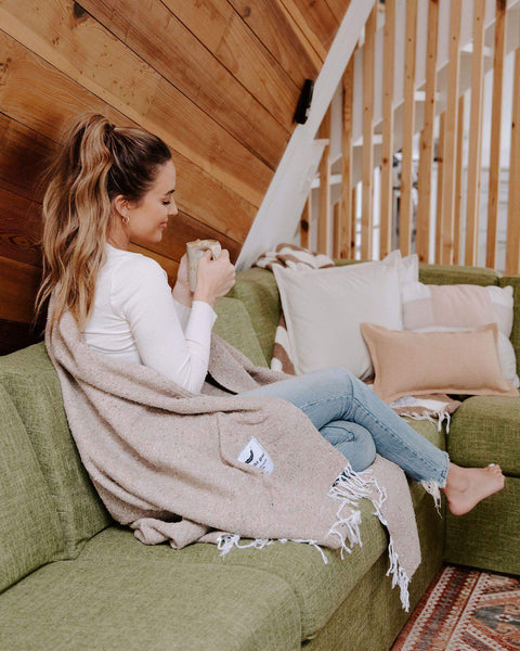 Cozy woman on green couch with Huntington Blanket, sand-colored and perfect for any adventure. Ideal for Trek Light outdoor fans.