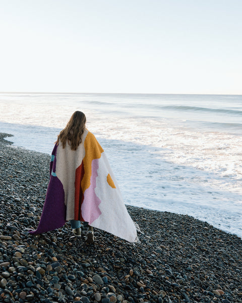 Cozy handwoven Mountain Sunrise Blanket made from recycled materials, draped over a person walking on a pebbled beach. By Trek Light.