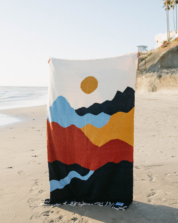 Colorful Trek Light blanket with mountain design draped on a sunny beach.