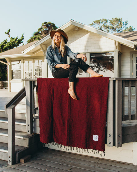 Woman in a hat sitting on a porch railing above a red Trek Light Napa Blanket, made from recycled materials. Eco-friendly cozy vibes.
