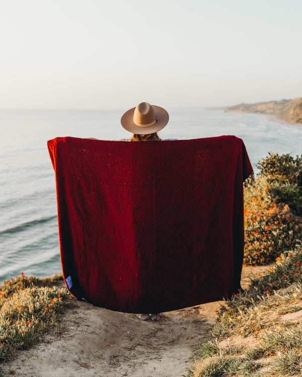 Person wrapped in red Napa Blanket by the ocean, cozy and eco-friendly, perfect for naps or enjoying nature. Trek Light.