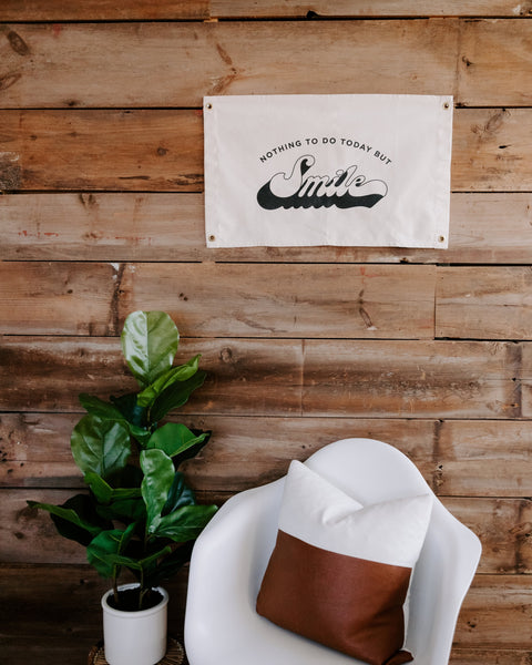Nothing To Do Today But Smile Canvas Flag, cream canvas with black print, hangs on a wood wall above a plant and chair.