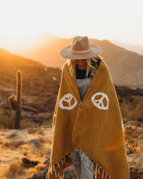Person wearing handwoven Peace Blanket from Trek Light in a scenic desert sunset, made from recycled clothing, cozy and beautiful.