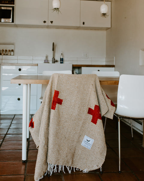 Trek Light Positive Vibrations Blanket draped over chair, handwoven with recycled materials, featuring red crosses and sustainable tag.