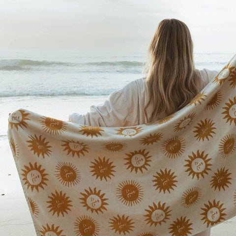 Woman on beach with Trek Light sunny bath towel made of sustainable cotton, showcasing sun patterns and super soft texture. Perfect relaxation.
