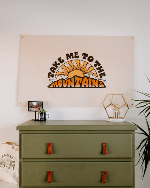 Take Me To The Mountains Canvas Flag in cream with a brown and orange sunburst print, hung above a green dresser with a lamp.