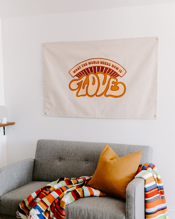What The World Needs Now Canvas Flag in natural canvas with retro orange LOVE print hangs above a gray sofa.