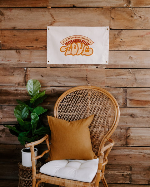 What The World Needs Now Canvas Flag in cream with orange LOVE print hangs on a wood wall above a rattan chair.
