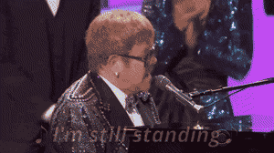 Elton John performing at the piano wearing a sparkling jacket during a live performance.