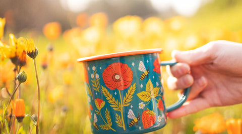 Hand holding a Trek Light Garden Buzz enamel mug with poppy and bee artwork in a field of orange wildflowers
