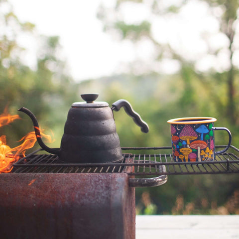 Midnight Mushroom enamel mug beside a black kettle at camp