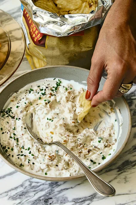 Hand dipping a chip into a bowl of sour cream and onion dip on a marble countertop
