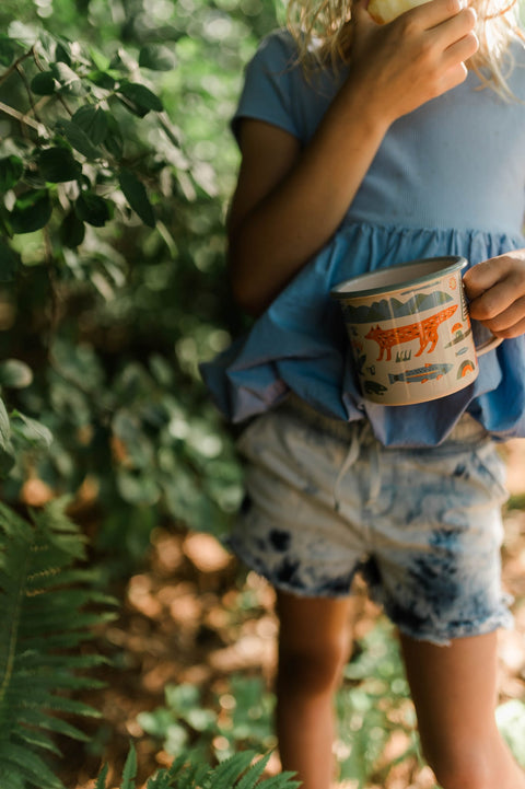 Person holding a Mountain Stream enamel mug 16oz outdoors surrounded by green foliage