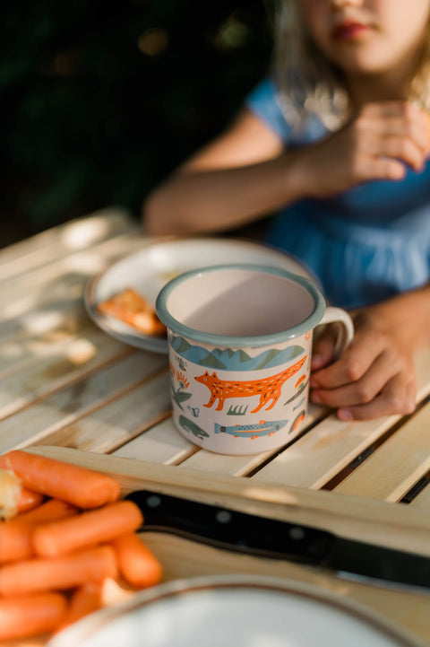 Mountain Stream enamel mug on a wooden picnic table with food at camp