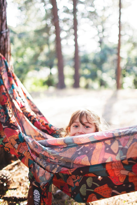 A child peeking out from a colorful Trek Light double hammock hung low in a forest.