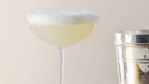 A refreshing cocktail in a coupe glass topped with frothy foam, next to a stainless steel shaker.