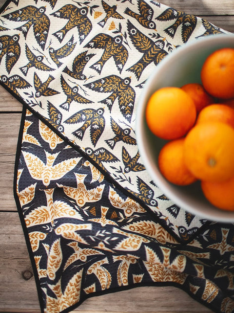 Songbird kitchen and tea towel laid on a wooden counter beside a bowl of oranges.