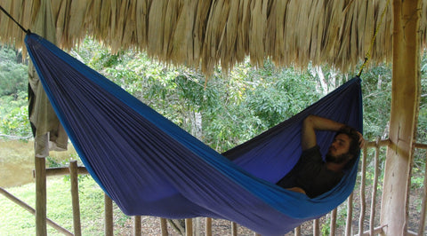 Man resting in a blue Trek Light hammock under a thatched roof shelter at the Nakum Maya site in Guatemala