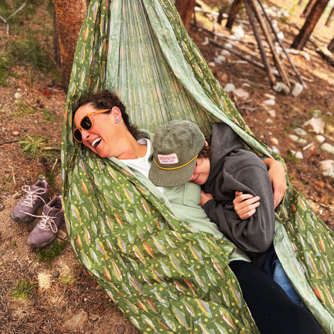 Two people relaxing in a Trek Light trout print hammock in a forest