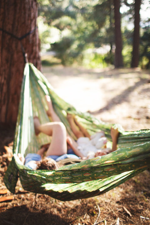 Two people relaxing in a Trek Light River Trout Print double hammock at a forest campsite
