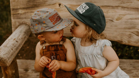 Two kids wearing caps, playfully eating watermelon slices, sitting on a wooden bench. Trek Light Kids Hats inspired playful moment.