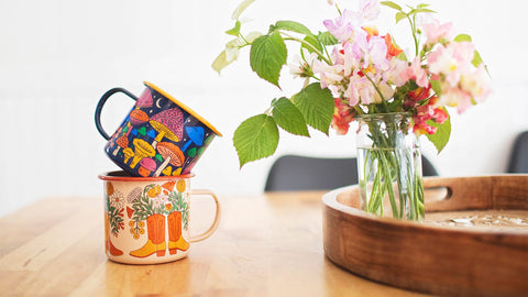 Colorful Trek Light enamel mugs stacked on a wooden table beside a vase of wildflowers.