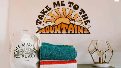 Sunset mountain wall art with a tote bag, colorful towels, and decorative lamp for a cozy Trek Light vibe.