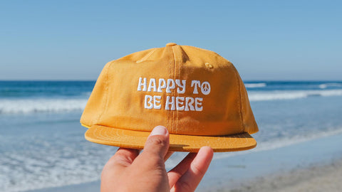 Trek Light Happiness Collection: Yellow cap with 'Happy to be Here' text, held by the beach, embodying joyful vibes.