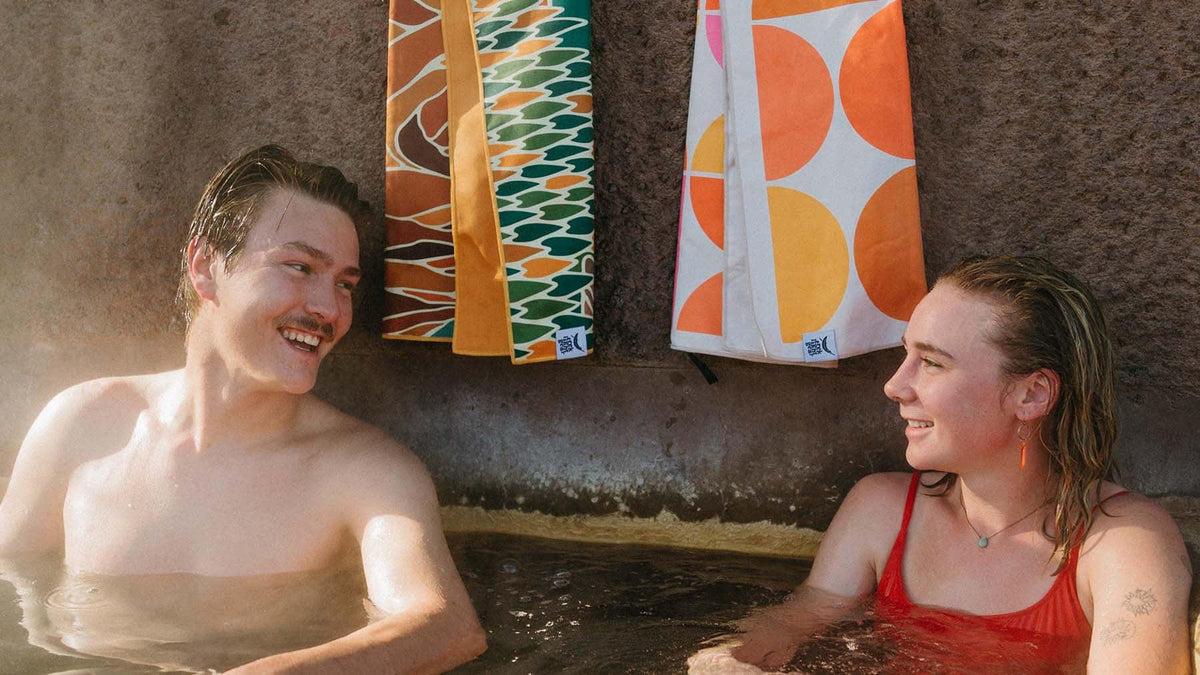 Couple relaxing with colorful Trek Light Wander Towels in background, super absorbent and eco-friendly travel towels.
