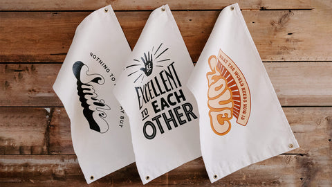 Three Trek Light canvas flags with inspiring messages hang on a rustic wooden wall, adding charm and positivity to your space.