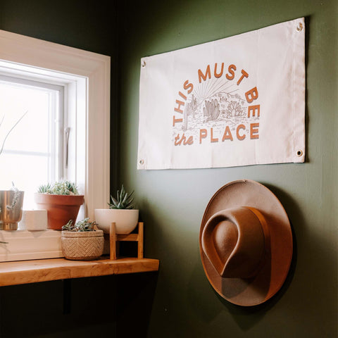 Cozy room with a 'This Must Be The Place' Trek Light canvas flag and cowboy hat hanging by the window, green wall accent.