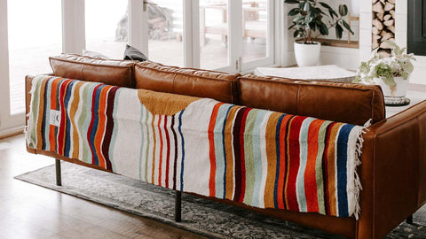 Colorful Trek Light blanket draped over a cozy leather sofa, adding charm to a stylish living room setup with home goods and decor.